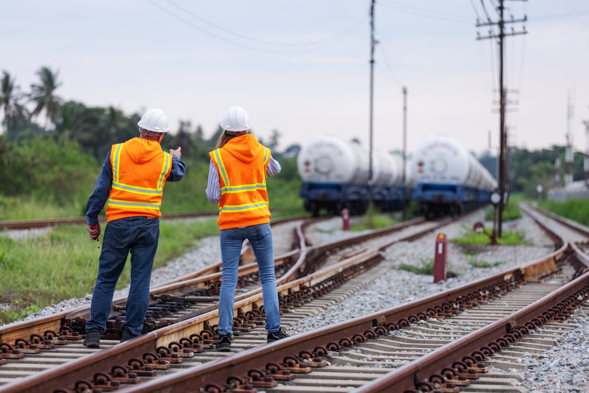Two railway workers wearing orange safety vests and helmets stand side by side on train tracks, observing a freight train in the distance, symbolizing teamwork, safety, and transportation oversight.